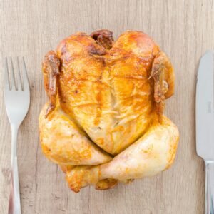 Top view of a perfectly roasted chicken placed between fork and knife on a wooden table.